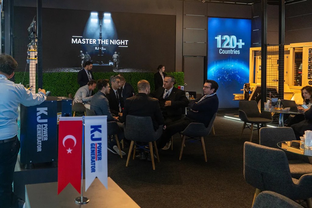 Bir etkinlik alanında toplanmış profesyonel erkek ve kadınlar, KJ Power Generator standında sohbet ediyorlar. Arka planda, "MASTER THE NIGHT" yazılı büyük bir ekran ve farklı ülkeleri temsil eden, mavi bir dünya haritası görüntüleniyor.