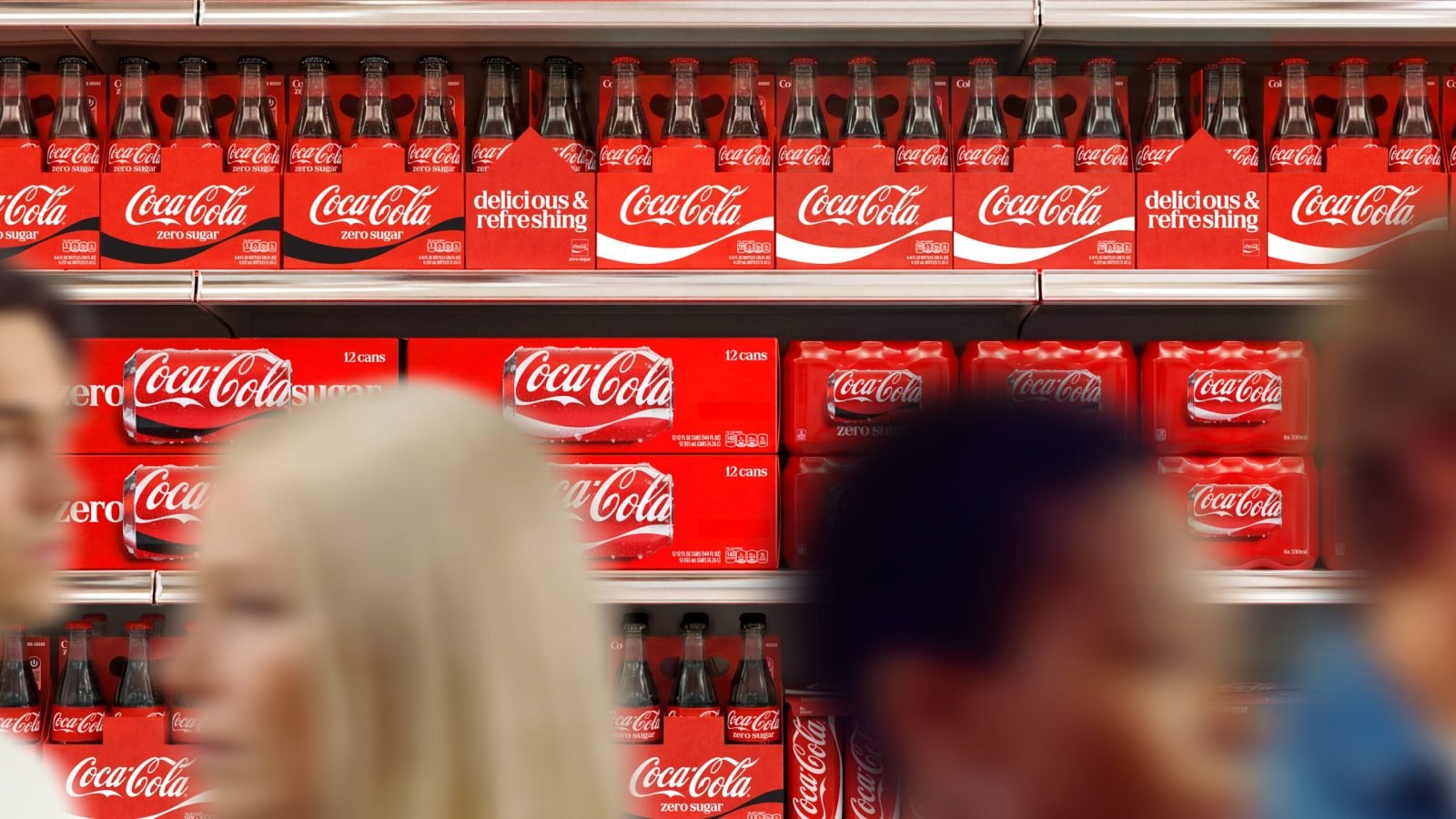 Market rafında dizilmiş Coca-Cola kolileri, önünden geçen bulanık insanlar