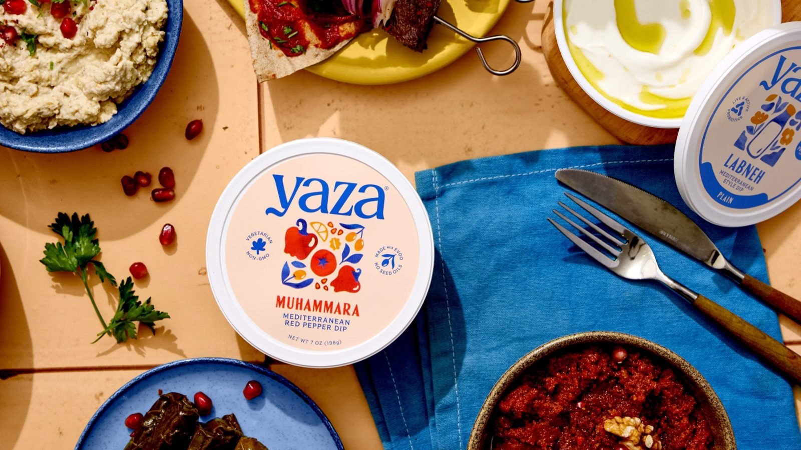 Üstten çekilmiş sofra: Yaza muhammara kabı, çeşitli mezeler, nar taneleri ve maydanozla dolu kaseler, mavi peçete ve çatal bıçak