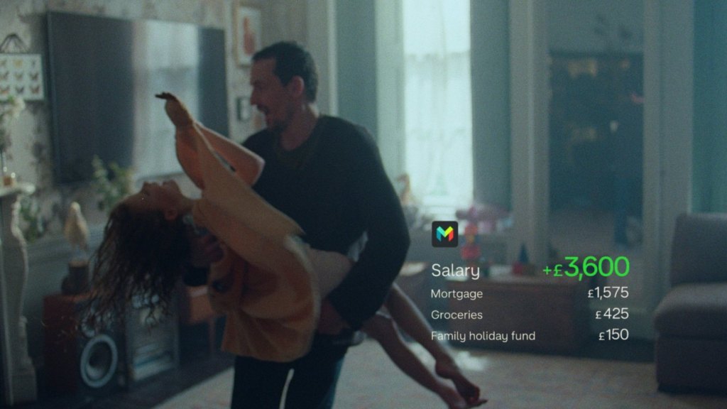Get busy living reklam filminden kare: oturma odasında dans eden bir çift, yanında Monzo uygulamasının maaş, kira, market ve tatil fonu satırlarını gösteren arayüz katmanı