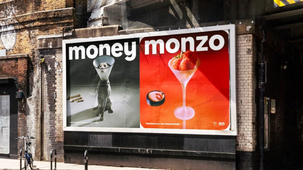Tuğla duvarda Money vs Monzo afişi: solda gri zeminde money yazısı ve yakalıklı köpek, sağda mercan kırmızısı zeminde büyük monzo logosu ve çilekli sorbe kadehi