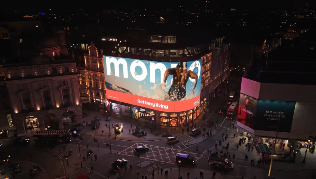 Londra Piccadilly Circus’ta gece, dev ekranda Monzo logosu ve Get busy living yazısı; meydanda trafik ve yayalar